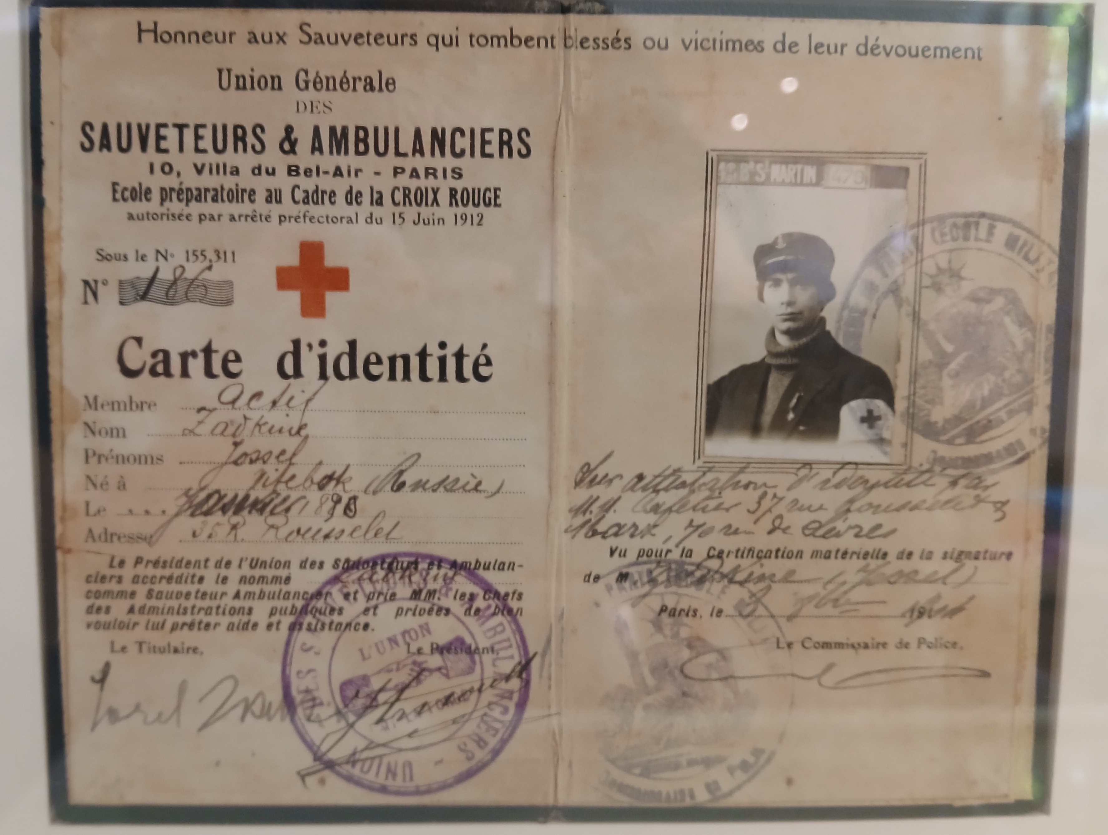 Document d'archive jauni en deux volets, avec une croix rouge, des mentions manuscrites, des tampons violets et la photographie d'un jeune homme en uniforme avec brassard.